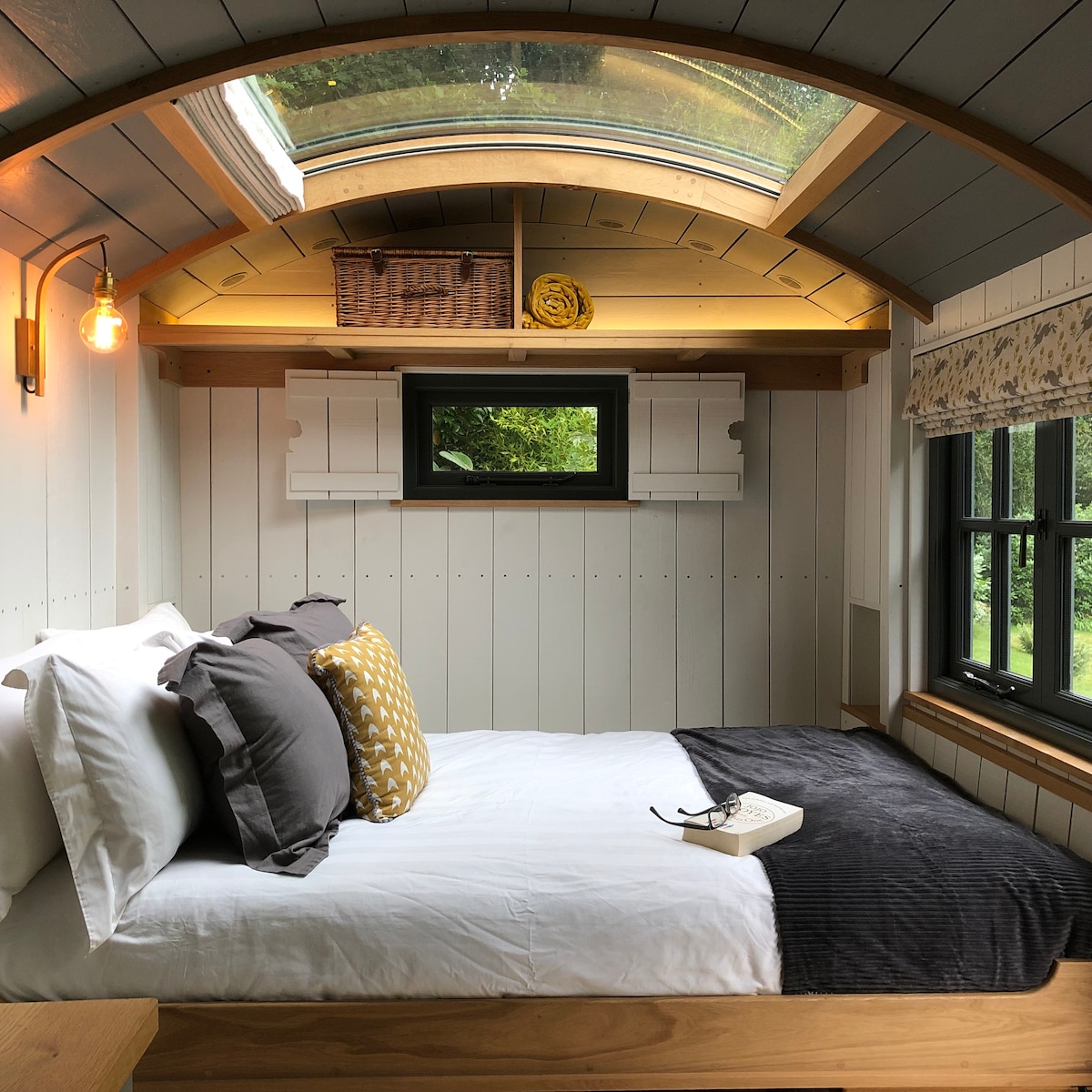 A cozy sleeping area features a dropdown queen bed adorned with a light-colored duvet, dark throw blanket, and decorative pillows. A glass ceiling star-gazing window allows natural light, while a shelf above holds a basket and rolled linens. Two windows framed in dark wood provide outside views.