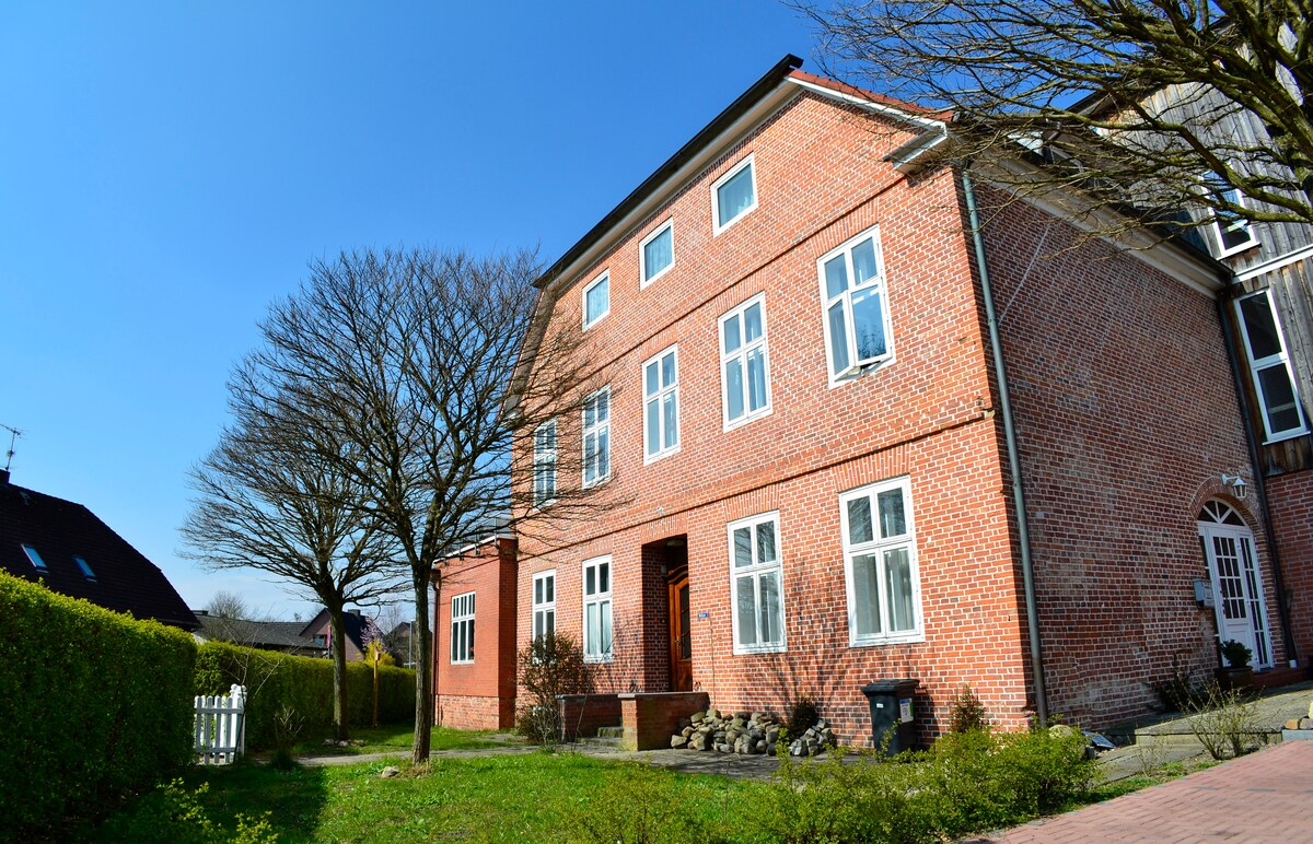A red brick building with multiple large windows is presented under a clear blue sky. Flanked by lush green hedges, the entrance features a small pathway leading to a door, complemented by a few stone decorations and a garden area.
