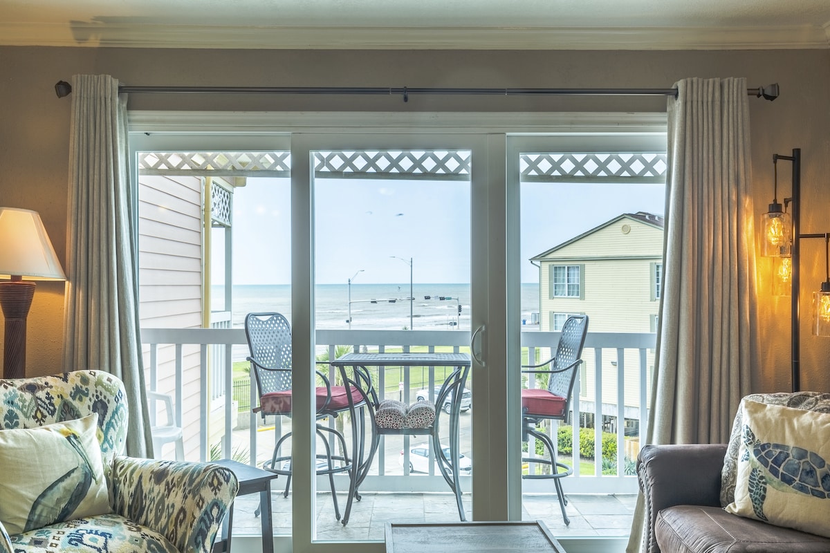 Floor-to-ceiling glass doors offer a panoramic view of the ocean, allowing natural light to fill the room. A cozy seating area features patterned chairs and a coffee table, while a private balcony with two chairs provides a space for relaxation and enjoyment of the sea breeze.