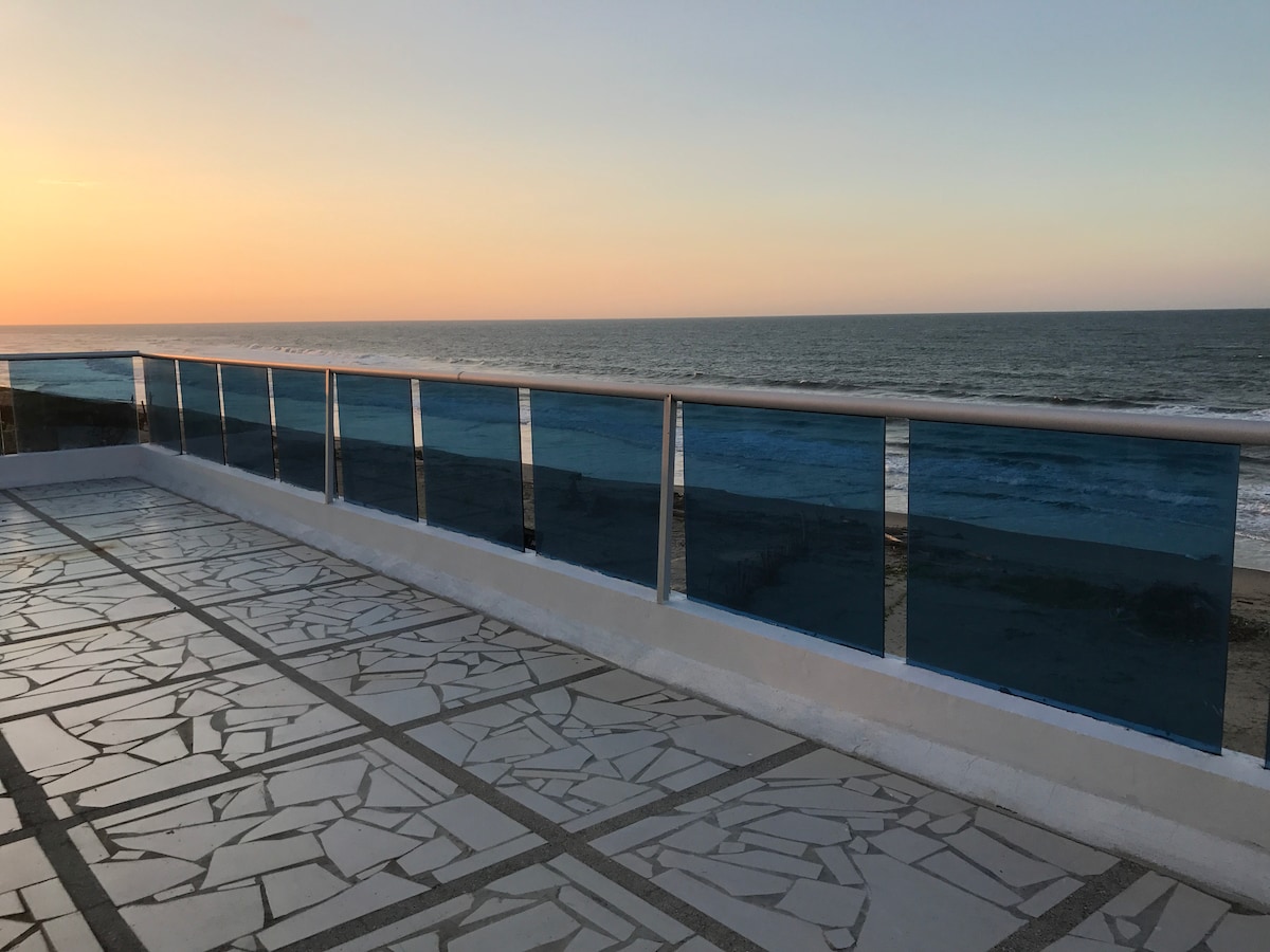 A spacious terrace is viewed, featuring a glass railing that overlooks the ocean. The floor is composed of patterned tiles, and a serene sunset casts gentle hues across the horizon. The sound of waves can be imagined from this inviting outdoor space.
