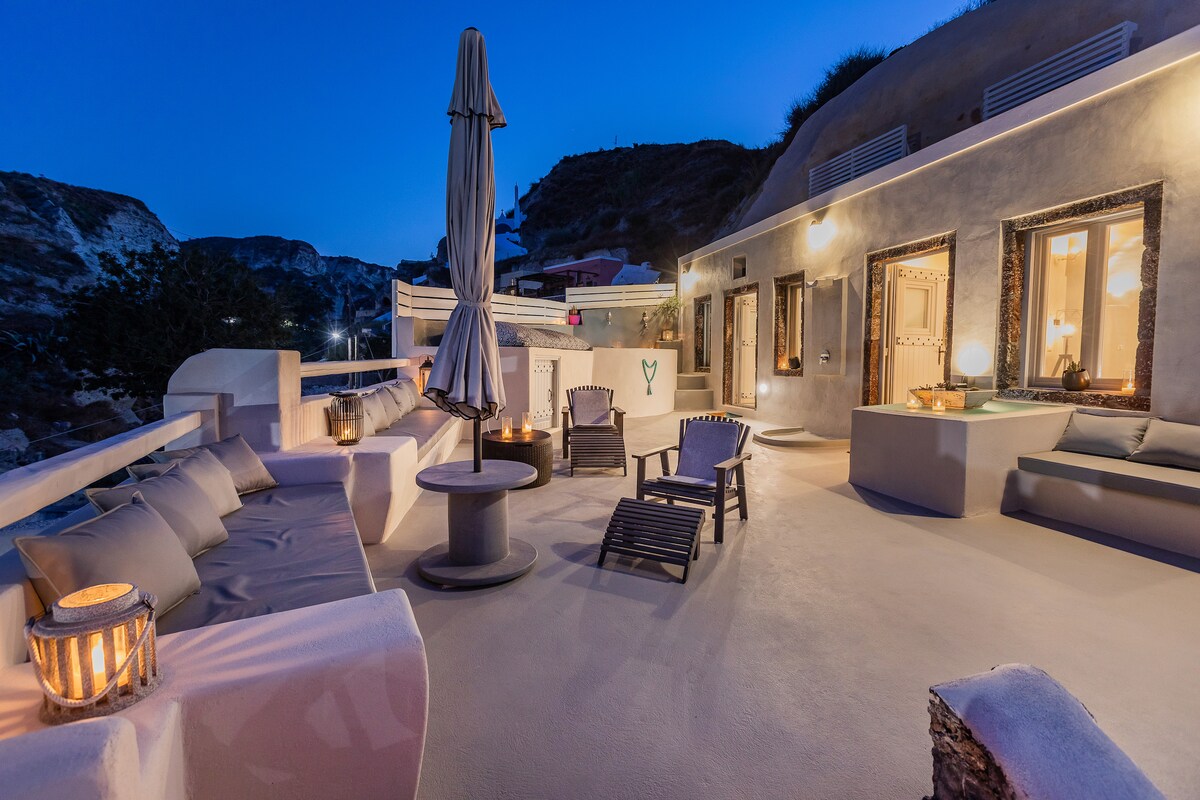 A spacious terrace is illuminated softly by lanterns, with a mix of seating including lounge chairs and a shaded area. The backdrop of rugged hills is visible, complemented by the charming facade of the villa. This setting provides a serene outdoor atmosphere.