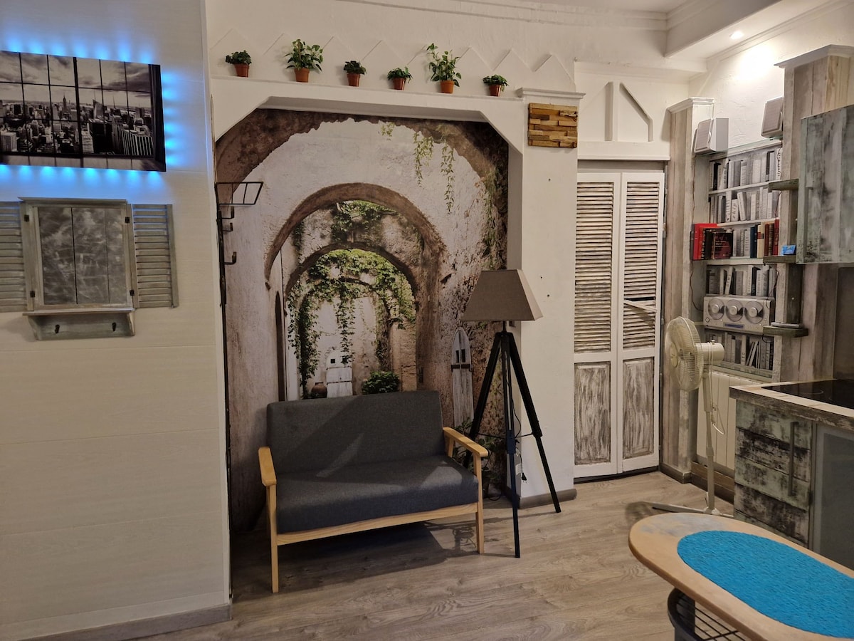 A cozy corner of the studio features a gray sofa in front of a wall adorned with an artistic mural of an archway draped in greenery. Shelves with books and decorative plants provide a refreshing touch, while a standing lamp adds gentle illumination.