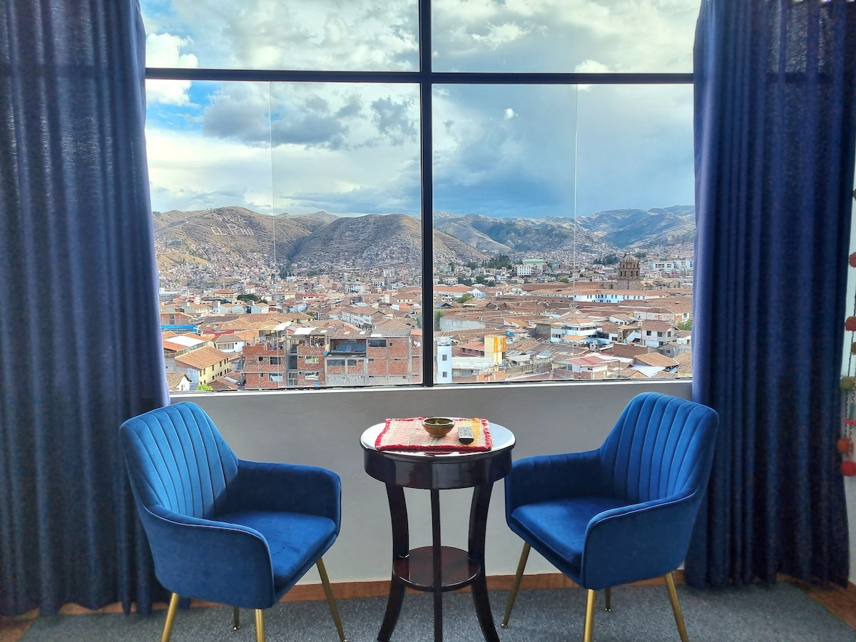 A pair of blue chairs are positioned beside a small table, offering a comfortable spot to enjoy panoramic views of Cusco. Large windows frame the vibrant cityscape and mountains in the distance, with flowing curtains adding a touch of elegance.