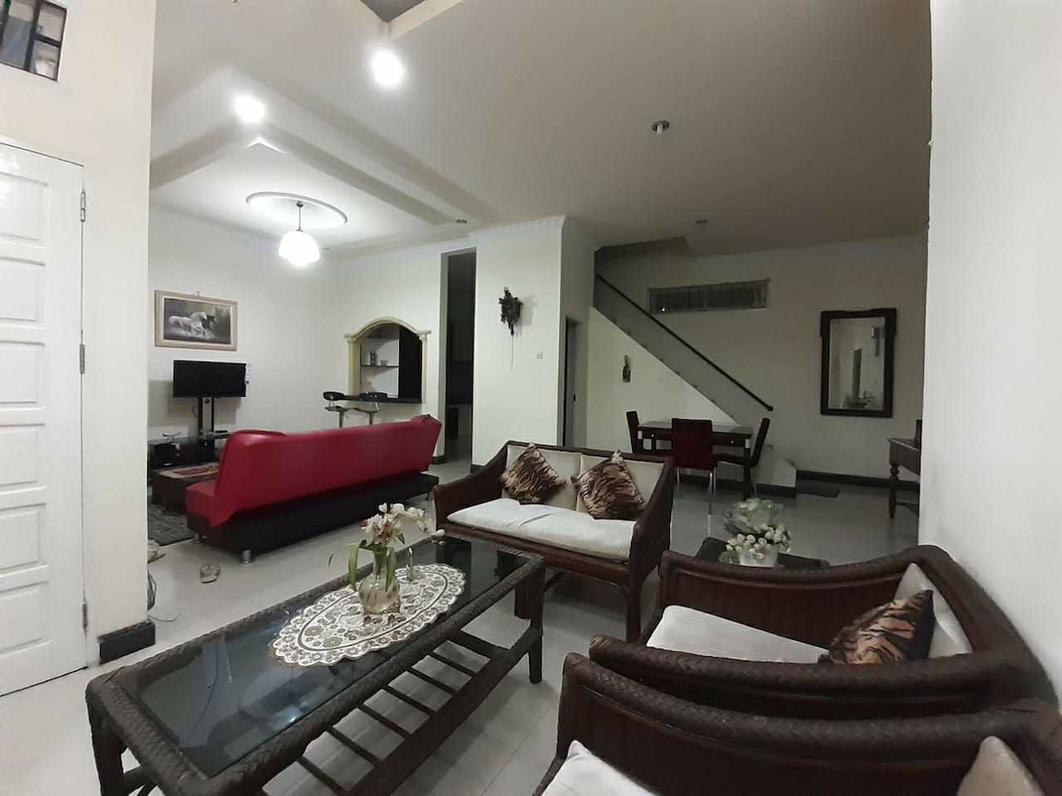 A welcoming living area features a comfortable red sofa and two wicker chairs surrounding a glass coffee table. Natural light fills the space, highlighting the minimalist decor and neutral-toned walls. A television is positioned for easy viewing, with a dining area visible in the background.
