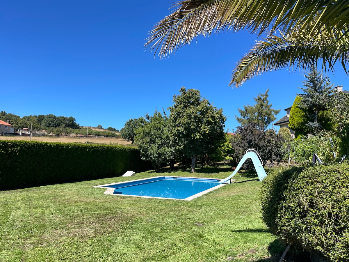 A clear blue pool is surrounded by well-maintained grass and lush hedges. An inviting waterslide curves into the pool, while trees provide shade in the background. The sky is bright blue, contributing to a cheerful outdoor environment.