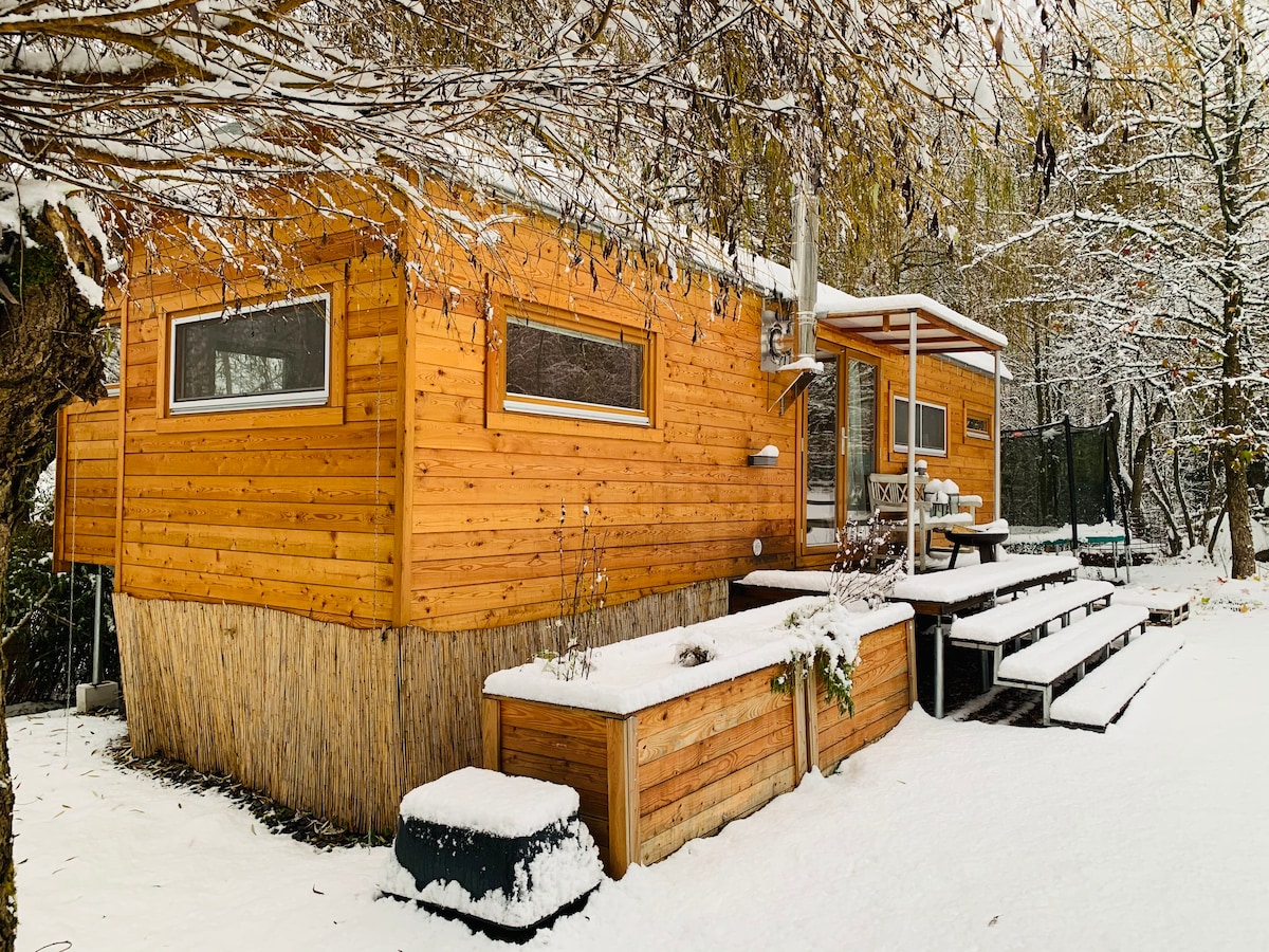 A cozy wooden retreat is surrounded by a blanket of snow, with a sloped roof and large windows reflecting the winter light. A set of wooden steps leads to a spacious outdoor deck, providing access to the natural landscape and nearby trees.