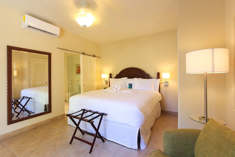 The interior of a guest room is displayed, featuring a spacious bed with a light-colored duvet. A large mirror reflects the room, which includes a bedside lamp and a small side table. Neutral tones are present in the decor, complemented by a green chair in the corner.