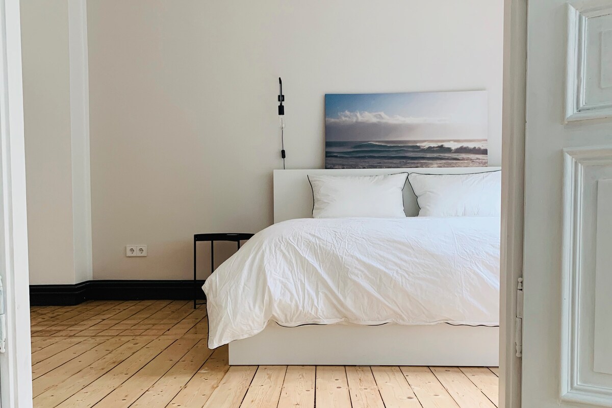 A serene bedroom is showcased with a spacious white bed complemented by soft bedding. A minimalist side table, positioned beside the bed, contrasts against the wooden floor. A large artwork featuring coastal imagery adorns the wall, contributing to a calming atmosphere.
