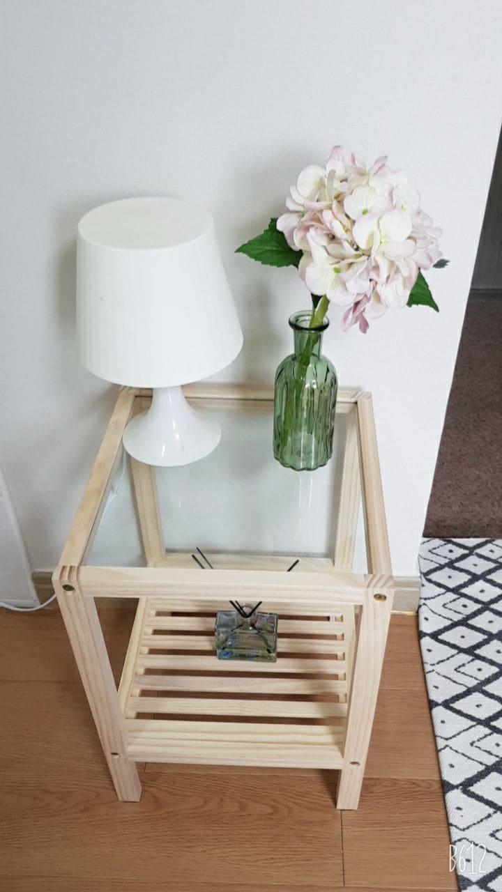 A modern side table is displayed with a glass top and wooden slats. It features a white lamp and a small vase containing a pink and white flower arrangement. The table's clean lines complement the minimalist decor.