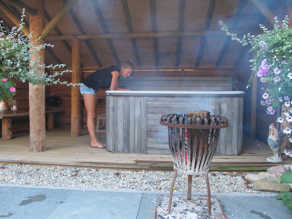 A rustic wooden sauna and jacuzzi area is surrounded by hanging flower pots, showcasing vibrant blooms. A small outdoor grill is positioned in the foreground, with embers glowing softly. The peaceful setting features a covered pavilion, allowing for year-round use.
