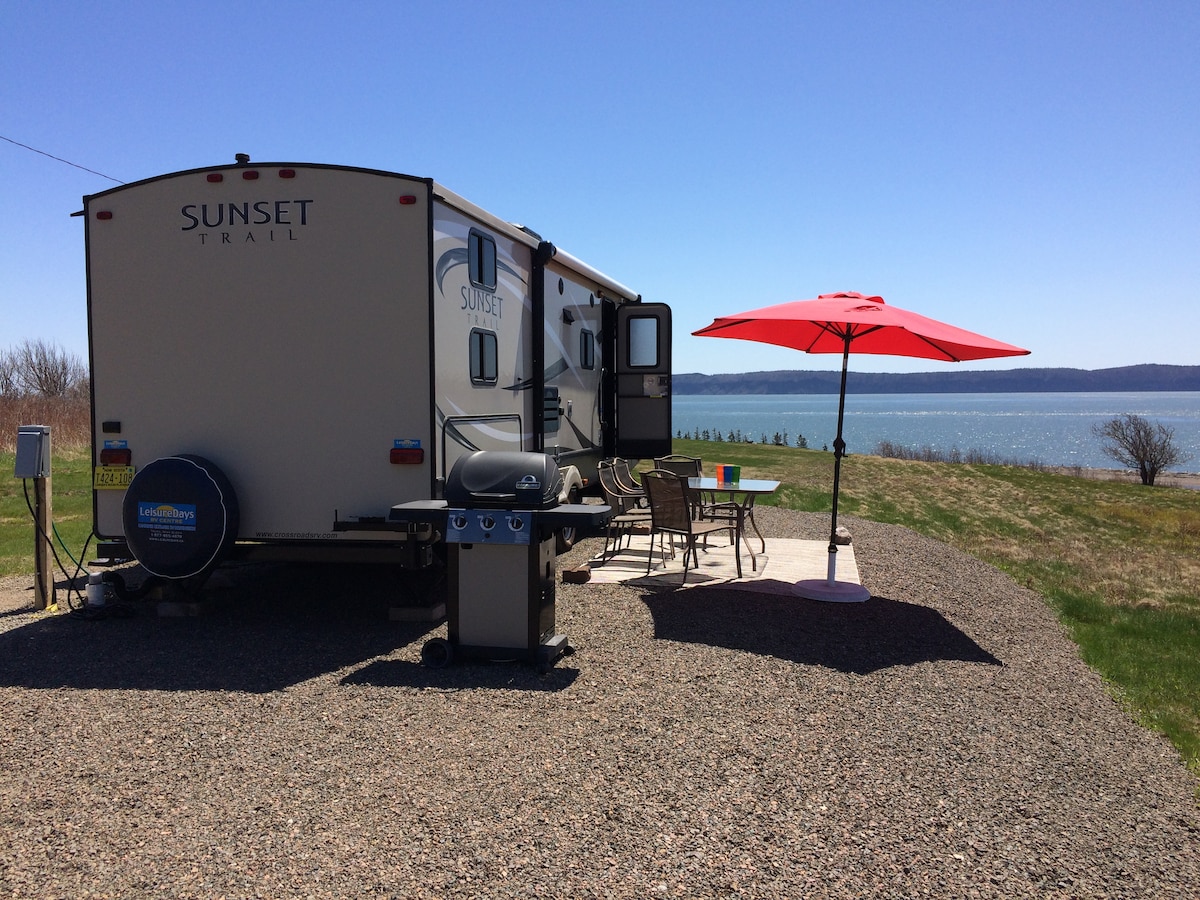 A cozy cottage travel trailer is positioned on a gravel patio, featuring a barbecue grill and a brightly colored outdoor dining table. A large red umbrella provides shade. The property overlooks a wide view of the ocean, with grassy areas extending toward the water.