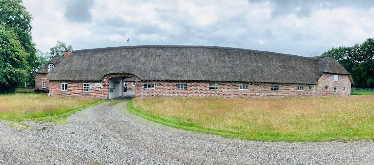 A scenic view of a traditional thatched-roof building, characterized by brick walls and a gravel pathway leading up to it. The surrounding area features lush green grass and a natural landscape, providing a serene setting typical of the region.