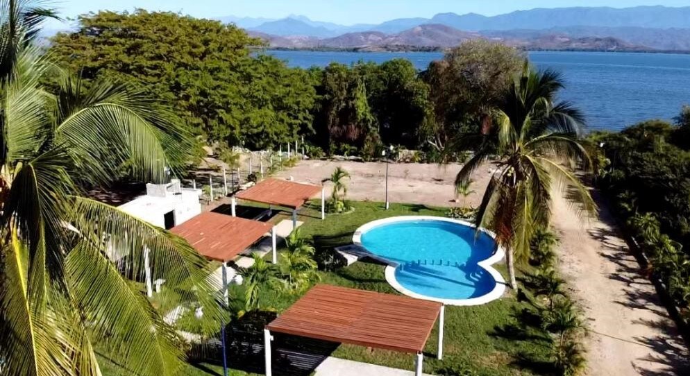 A serene outdoor space features a swimming pool surrounded by lush greenery and palm trees. Several shaded seating areas with wooden pergolas are positioned nearby. The tranquil waters of the lagoon reflect the mountains in the background, providing a scenic view of the natural landscape.