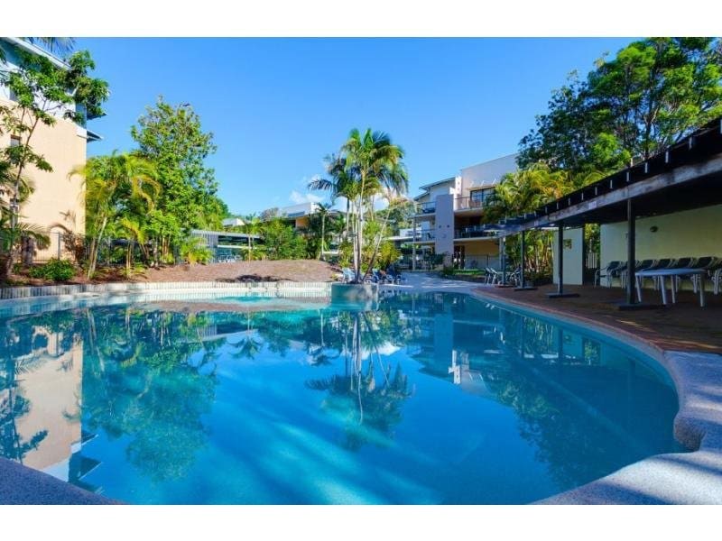 The lagoon pool is surrounded by tropical landscaping, with clear blue water reflecting the sunlight. Sun loungers are positioned along the deck, providing a relaxing space. The resort buildings are visible in the background, adding to the serene environment.