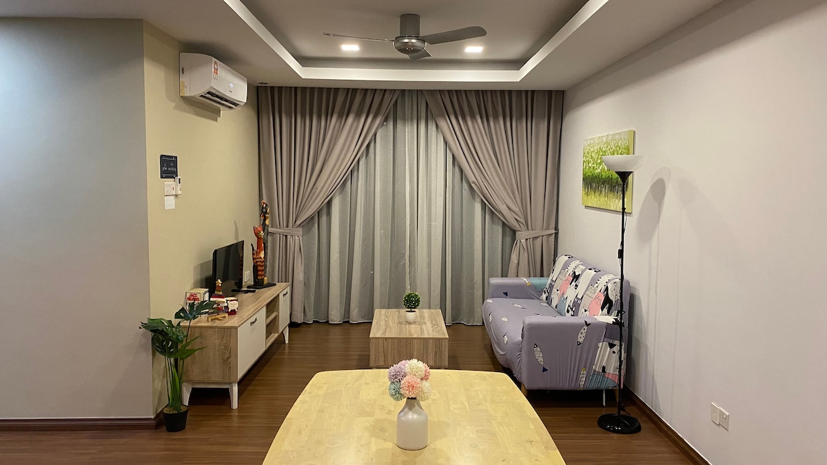 The living room features a light purple sofa with decorative cushions positioned near a wooden coffee table. A TV console is visible under the wall-mounted television. Sheer curtains gracefully frame a large window, complemented by a ceiling fan and a standing lamp in the corner.
