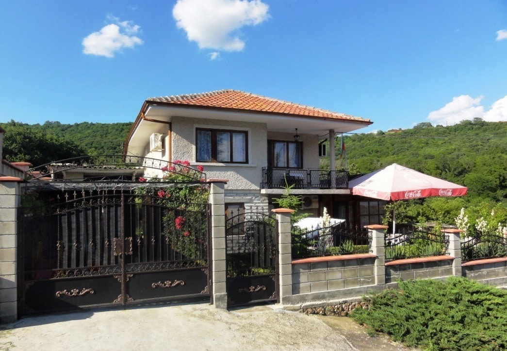 A two-story villa is surrounded by lush greenery, featuring a tiled roof and a decorative metal gate. A shaded outdoor seating area is visible, offering a cozy spot in front of the building. The tranquil landscape adds to the serene ambiance of the property.