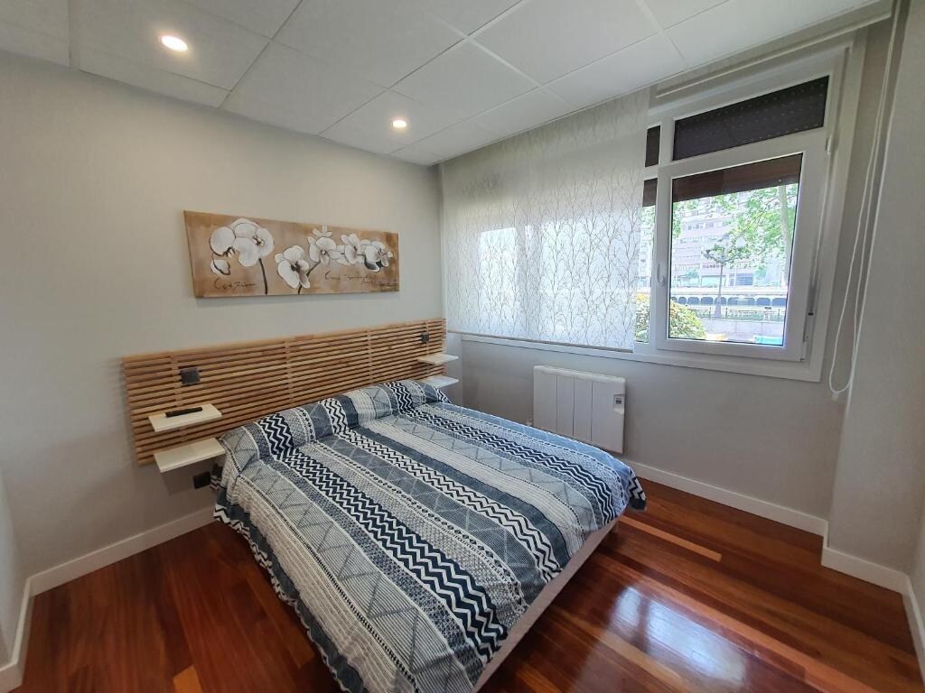 A spacious sleeping area features a patterned bedspread with blue and white stripes atop a generous bed. Wooden headboards are mounted on either side, equipped with bedside tables and lamps. Bright light floods the room from large windows overlooking greenery outside.