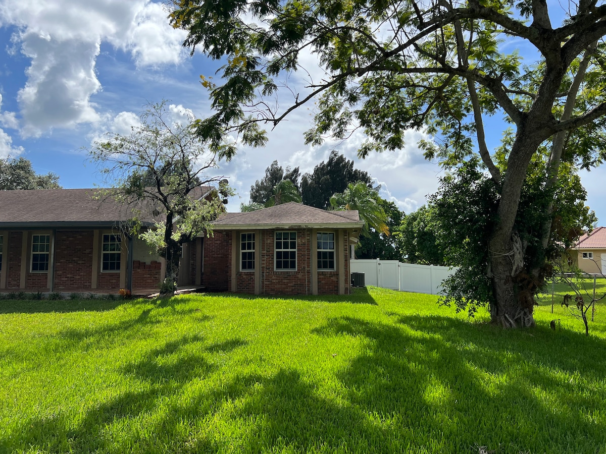 A private guesthouse is surrounded by lush greenery, featuring a well-maintained lawn. Tall trees cast gentle shadows across the vibrant grass, while the building is set against a backdrop of partly cloudy blue skies.