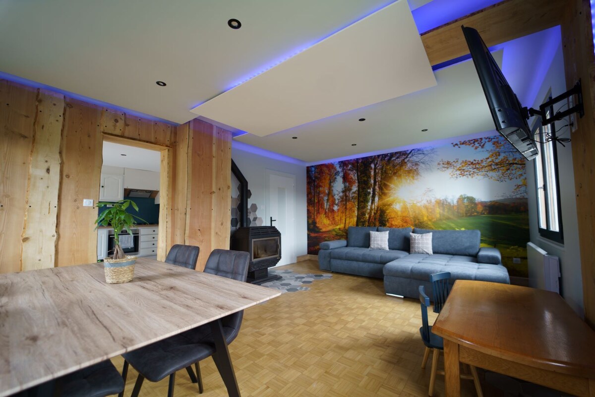 The inviting living area features a spacious sectional sofa, a dining table with dark chairs, and a carefully placed flat-screen TV. Soft lighting creates a comfortable ambiance. A large mural of autumn trees adds a touch of nature to the wooden walls.