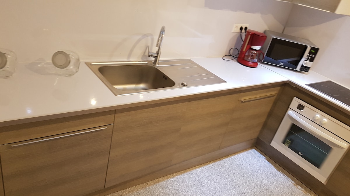 A modern kitchen is shown, featuring a stainless steel sink integrated into a polished countertop. Cabinets below provide storage space, while a microwave, coffee maker, and oven are visible on the countertop, highlighting the kitchen's functionality and design.
