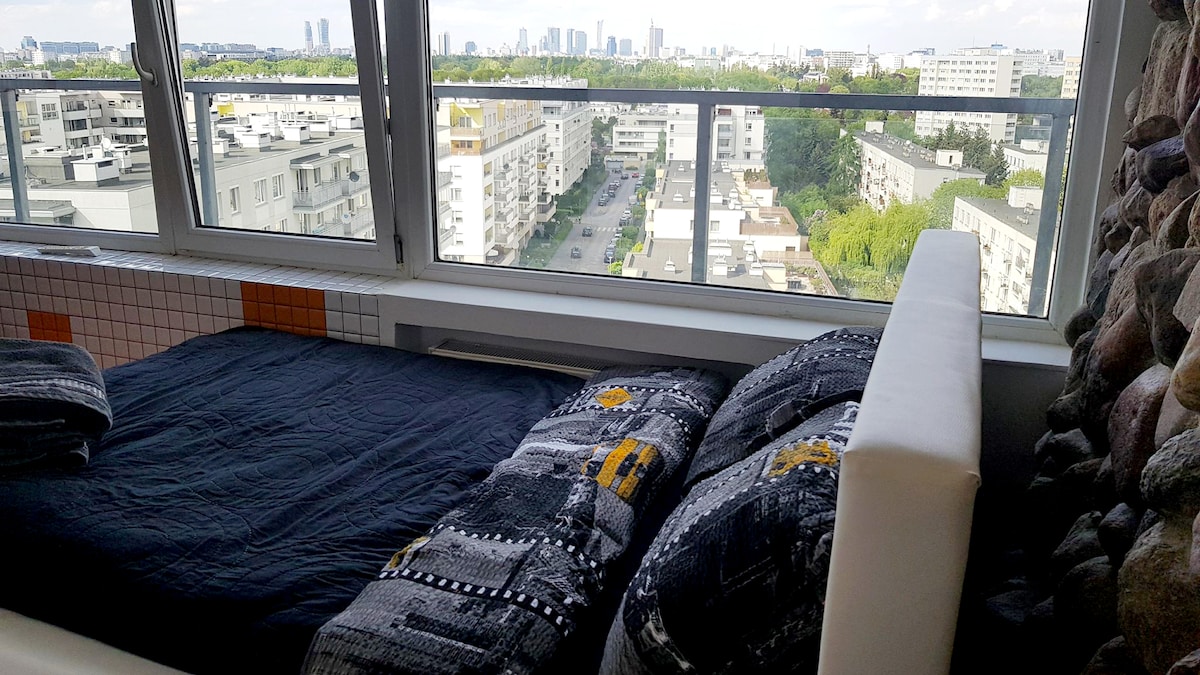 A spacious bed is positioned near large windows, offering expansive views of the city skyline and surrounding green areas. The bedding features a modern design with vibrant patterns, and natural light fills the room, enhancing the serene atmosphere.
