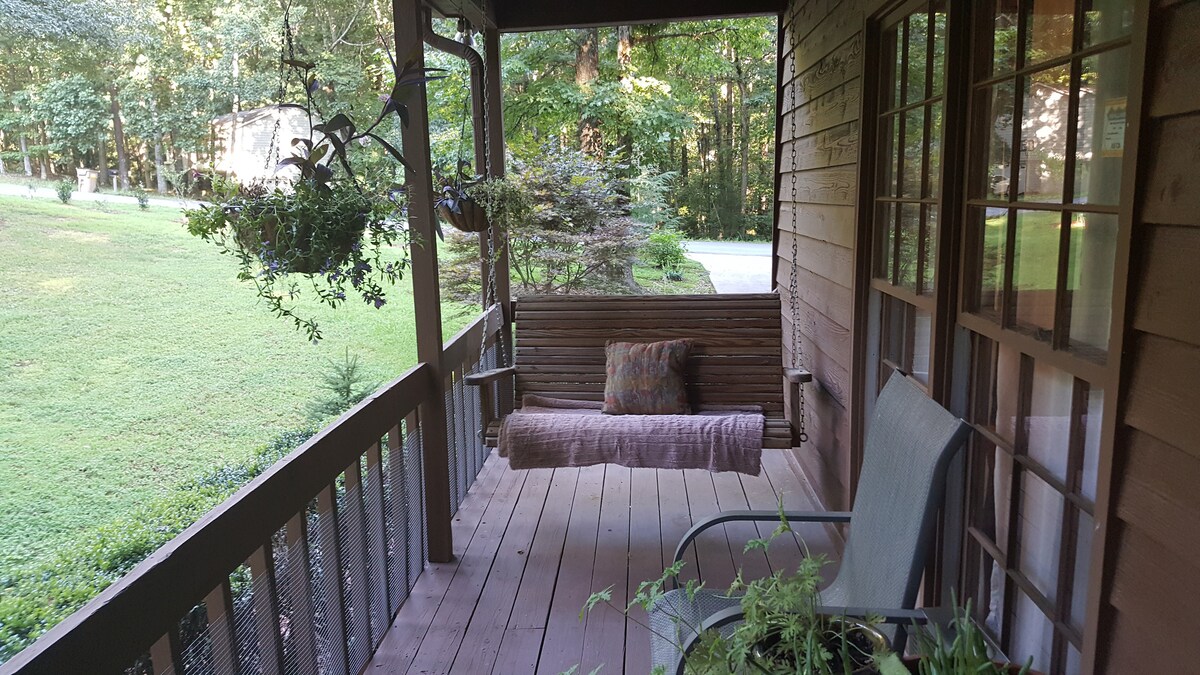 A front porch is depicted with a wooden swing and a small side chair. Lush greenery surrounds the space, while hanging plants add a touch of nature to the entryway. The floor is crafted from wooden planks, inviting relaxation and enjoyment of the outdoor view.