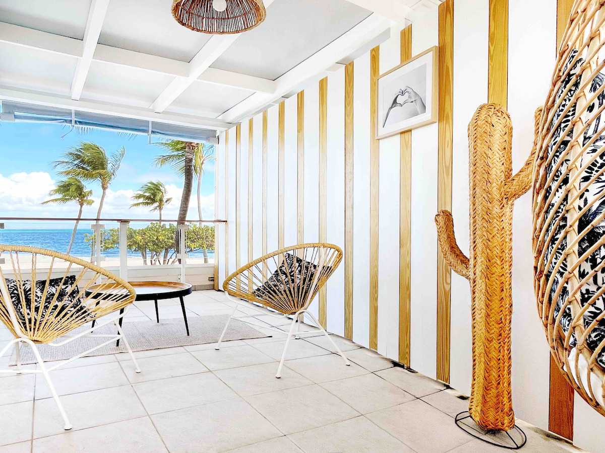 A bright seating area is highlighted by large windows offering a view of the beach and palm trees. Two woven chairs surround a low coffee table, while a decorative woven cactus enhances the coastal theme. Light colors and wood accents create a welcoming atmosphere.
