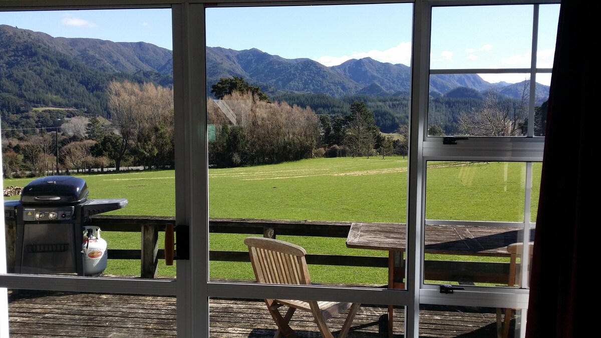 A panoramic view showcases lush green fields and distant mountains beyond a wooden deck. A barbecue grill and a wooden table with a chair are positioned on the deck, allowing for outdoor relaxation amidst the tranquil landscape.