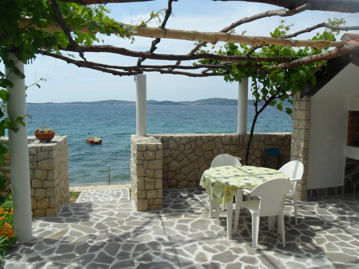 A serene outdoor seating area is present, featuring a table adorned with a floral tablecloth and four white chairs. The space is shaded by natural vines and offers unobstructed views of the sea and surrounding landscape.