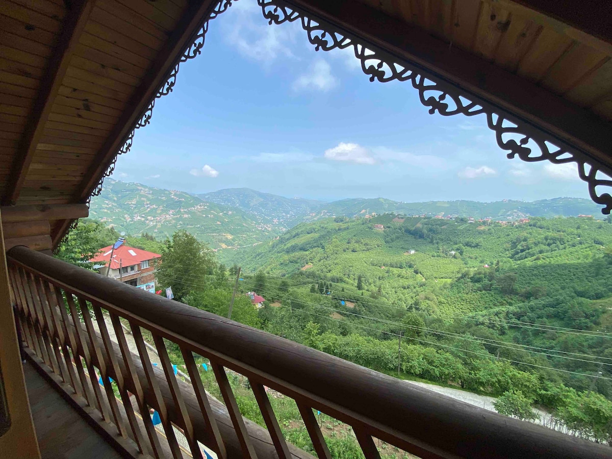A wooden balcony offers a serene view of rolling green hills and valleys. Lush trees and distant mountains are visible under a partly cloudy sky. The natural landscape creates a peaceful backdrop, inviting guests to relax in the fresh air.