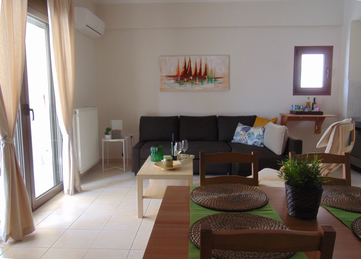 A spacious living area features a comfortable dark sofa and a light wooden coffee table, accompanied by green decorative elements. Natural light streams through large glass doors that open to a balcony. A dining table set for four is positioned nearby, complemented by a subtle artwork on the wall.