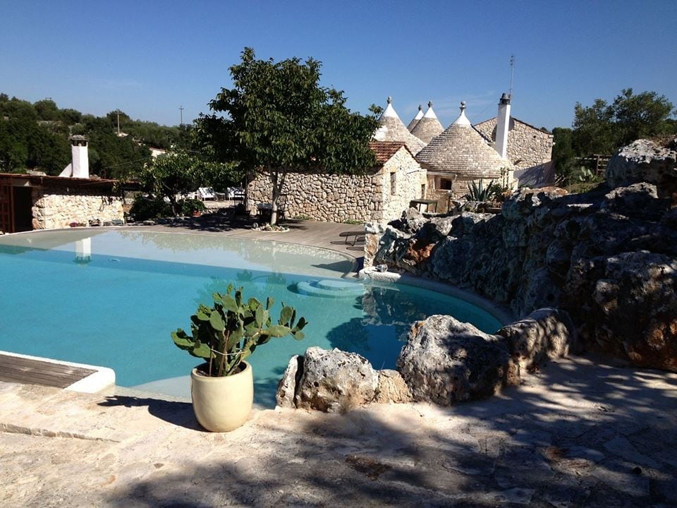 The image features a serene stone-pool area surrounded by natural rock formations and lush greenery. Gentle ripples can be seen on the water's surface. Traditional trulli structures are visible in the background, showcasing iconic conical roofs under a clear blue sky.