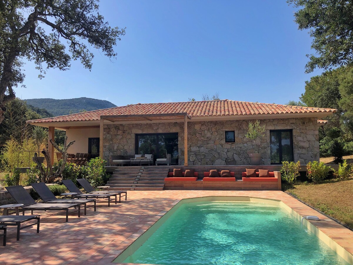 The villa features a rustic stone exterior with a terracotta roof, surrounded by lush greenery. A private pool with sun loungers is positioned in the foreground, offering a relaxing outdoor space. Large glass windows invite natural light into the living area, connecting interior and exterior.