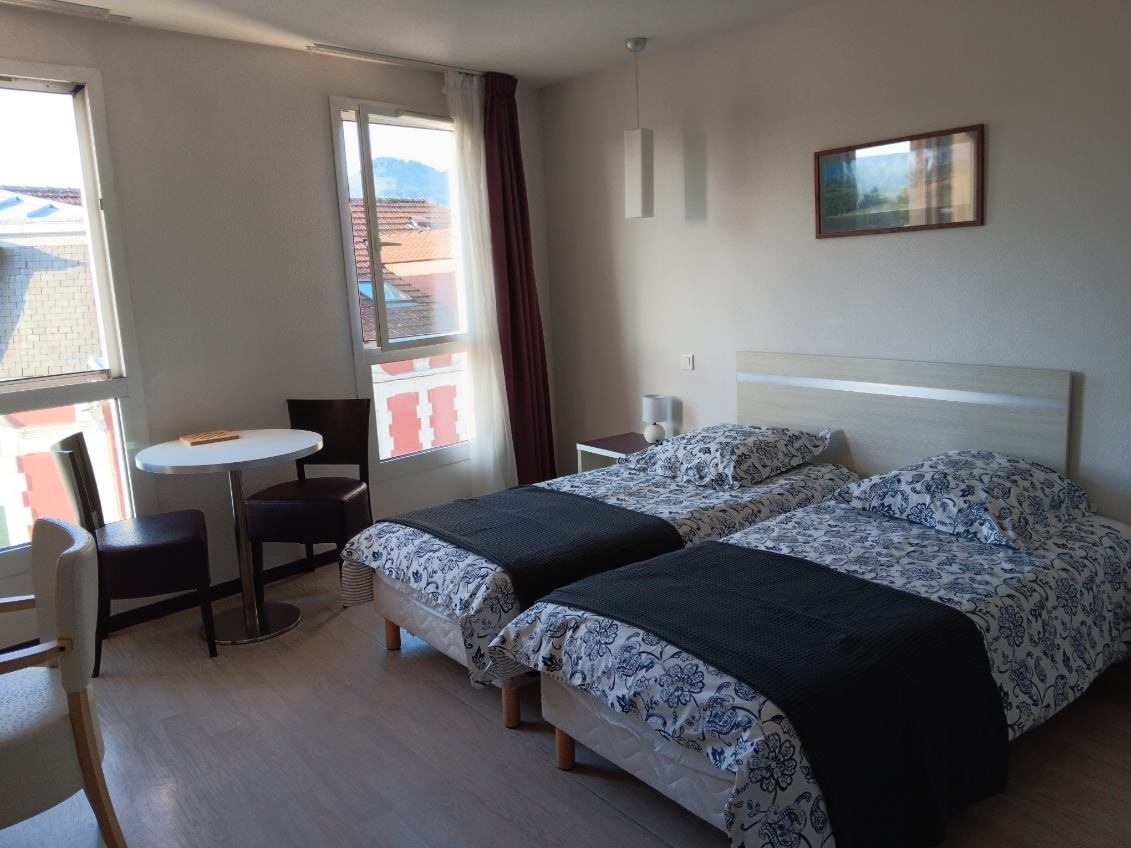 The bedroom features two single beds with patterned linens separated by a dark blanket. A small table with two chairs is positioned near large windows, allowing natural light to fill the space. A simple light fixture and wall art add subtle decor.