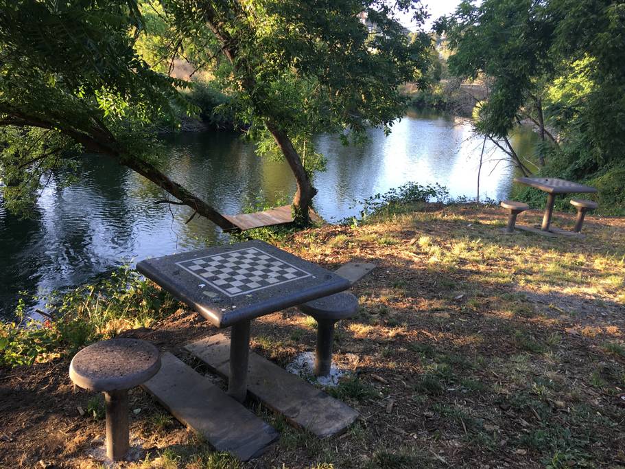 A chess table with two stone benches is positioned near the riverbank, offering a scenic view of the calm water and lush greenery. The surrounding area features natural foliage, providing a tranquil setting for outdoor activities.