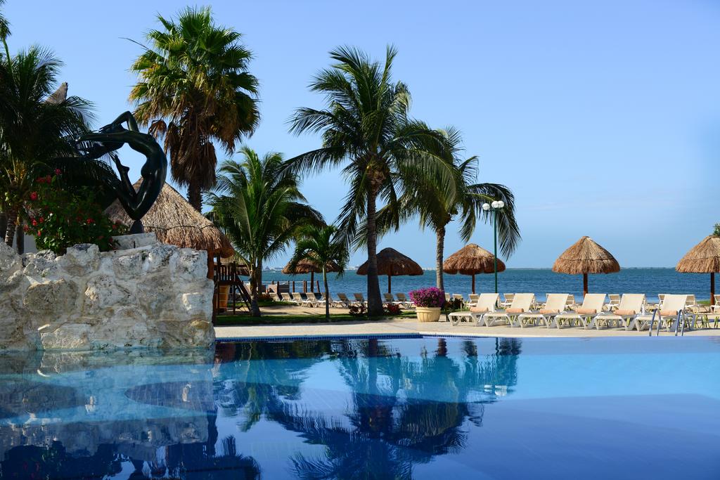 A refreshing pool reflects the clear sky and nearby palm trees, with thatched umbrellas providing shade along the beach. Sun loungers are positioned for relaxation near the water's edge, while vibrant flowers add a pop of color to the serene environment.