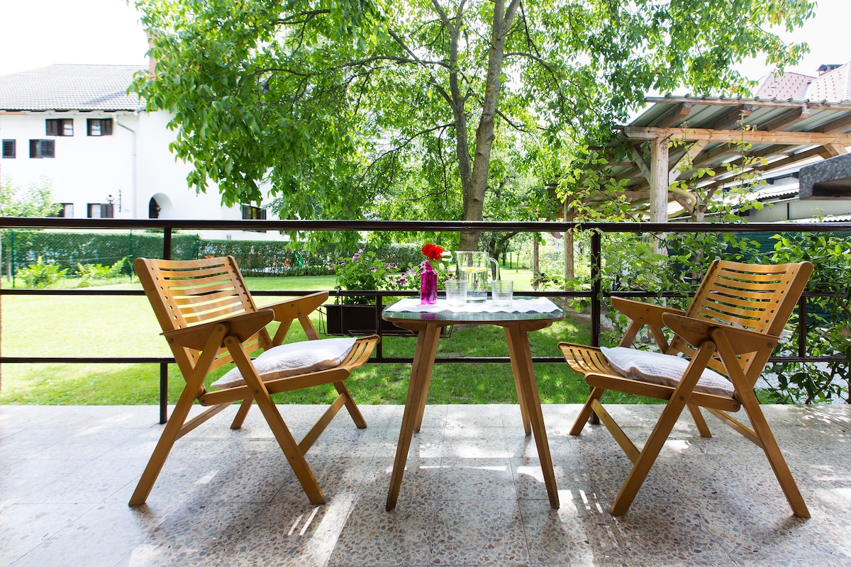 A serene outdoor terrace features two wooden chairs and a round table adorned with a vase of flowers. Lush greenery surrounds the area, providing a peaceful view of the garden and inviting relaxation in a tranquil setting.