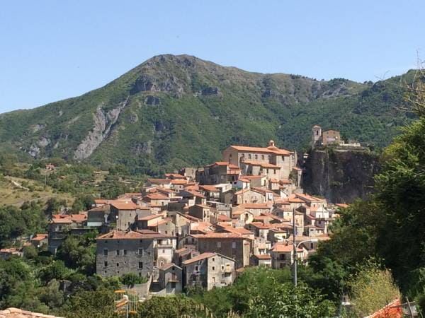 A scenic view of Papasidero is captured, showing a cluster of stone buildings with terracotta roofs nestled against a mountainous backdrop. The landscape features lush greenery and rocky slopes, highlighting the natural beauty surrounding this historic town.