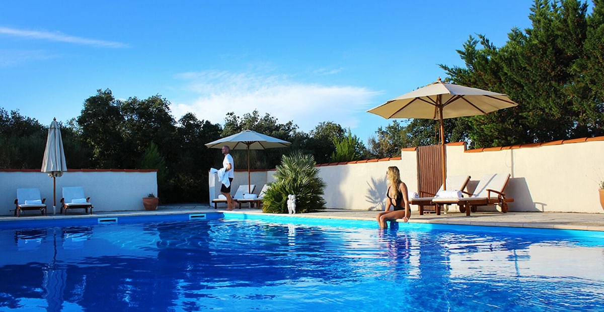 A serene swimming pool is surrounded by lush greenery and features comfortable lounge chairs under shaded umbrellas. The clear blue water reflects the sky, providing a refreshing atmosphere for relaxation and enjoyment.
