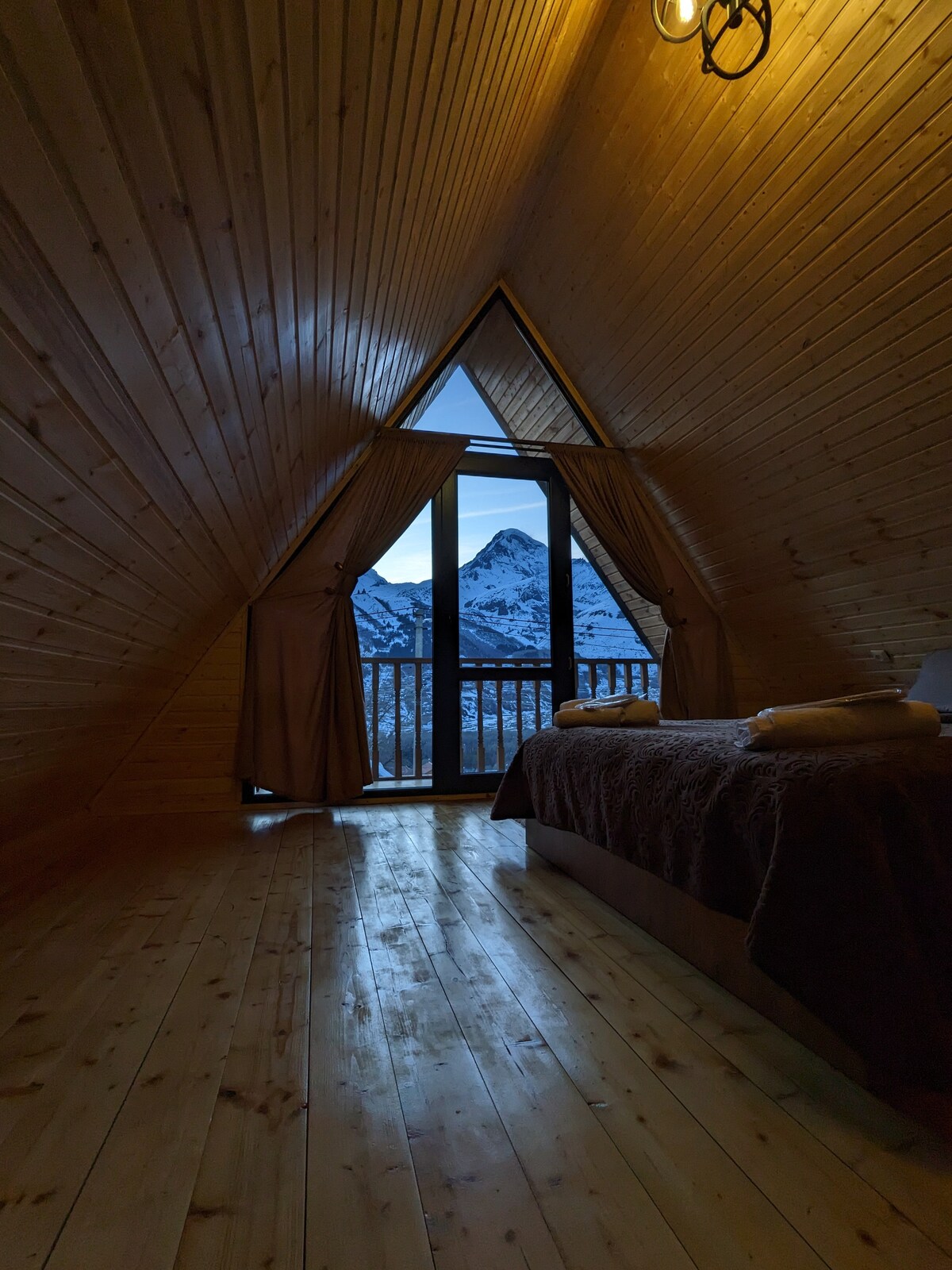 A triangular room features wooden paneling and flooring, creating a warm ambiance. A spacious bed is positioned beside a large window, offering a view of the mountains. Soft curtains frame the window, while folded towels rest on the bed, adding to the cozy setting.