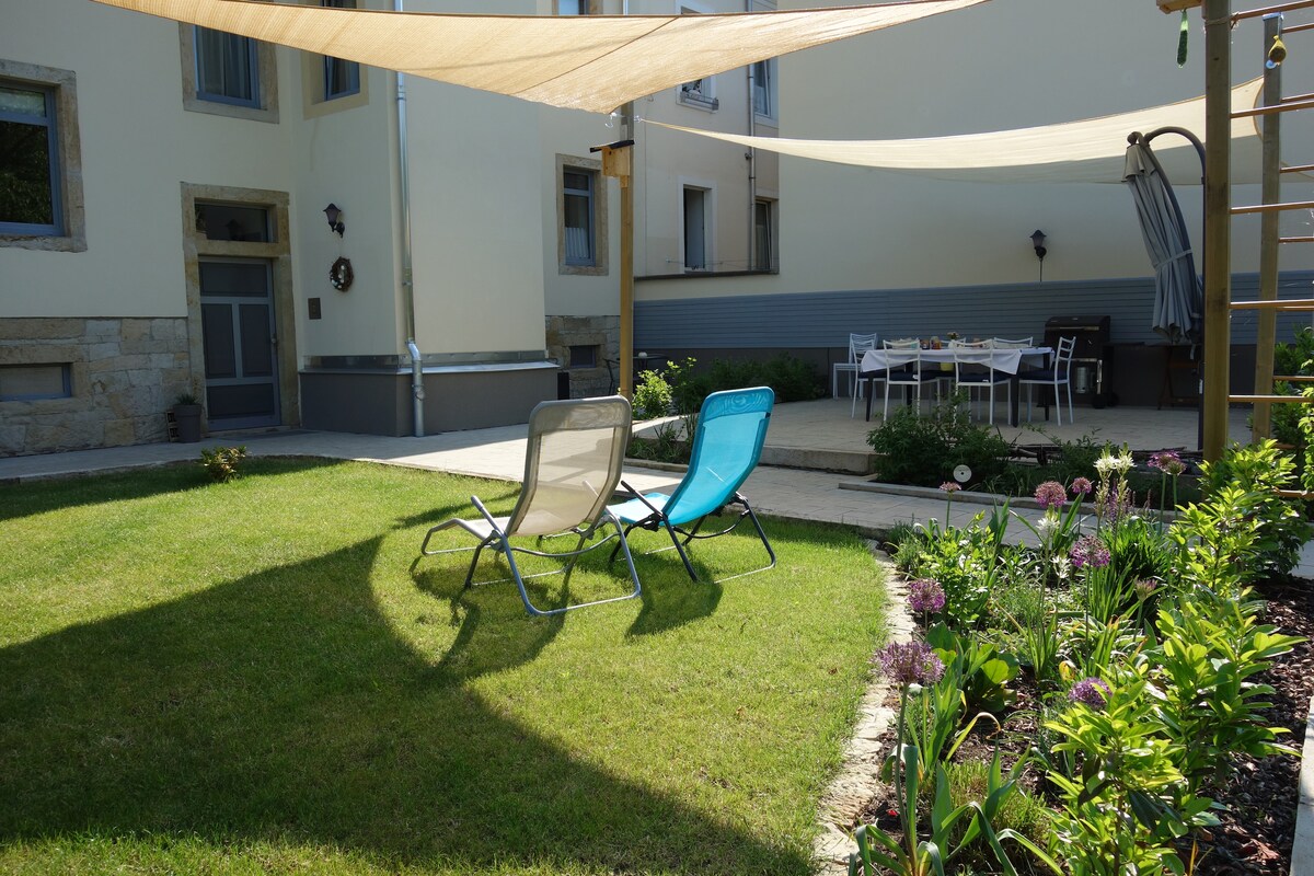 A well-maintained garden area features two lounge chairs set on a neatly trimmed lawn. Sunshade sails provide shelter over a dining table, visible in the background. Flowerbeds with colorful plants border the space, contributing to a peaceful outdoor environment.