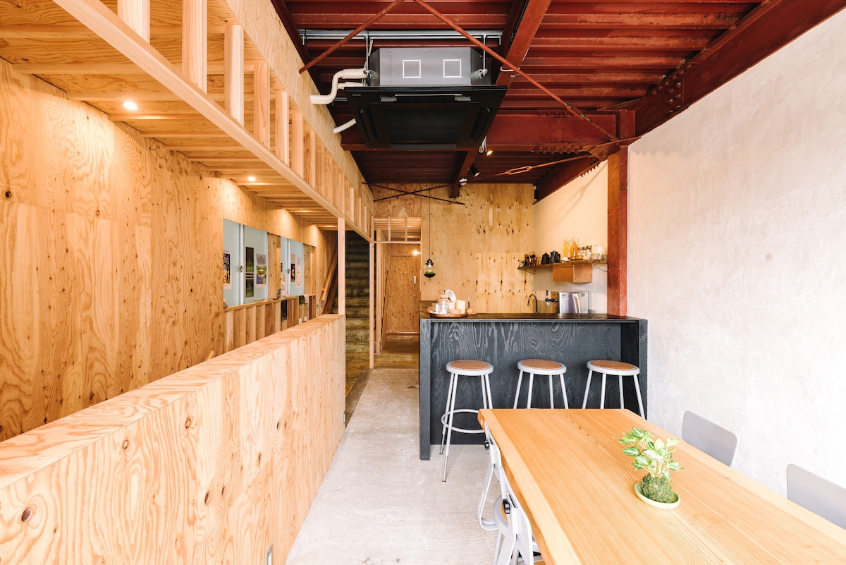 An open communal space is highlighted by natural wood paneling and understated decor. A kitchen area features a black counter with seating for three, while a dining table for six is positioned nearby. Subtle lighting and open architecture contribute to a welcoming ambiance.