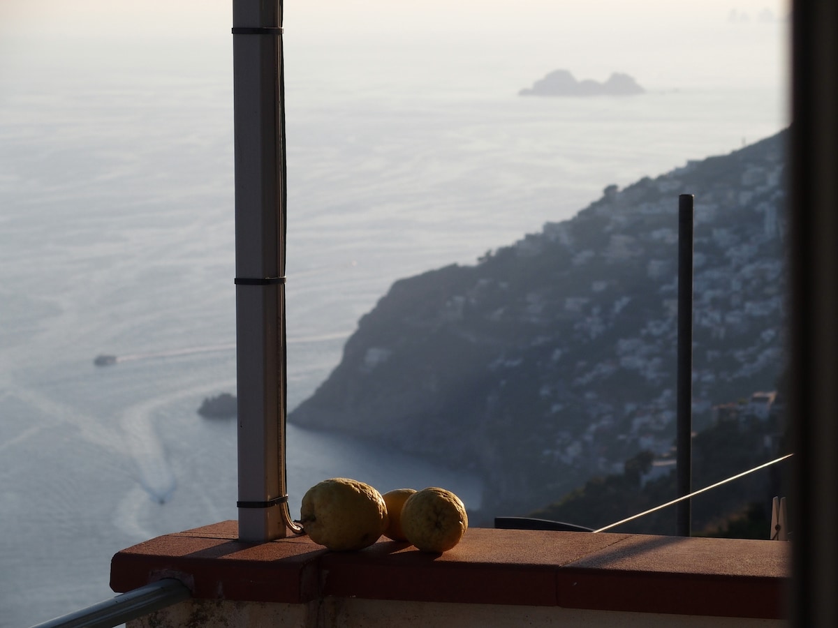A balcony railing is visible, adorned with two round fruits. In the background, a hazy view of the coastline can be seen, with the shimmering sea reflecting the light. Islands are faintly discernible on the horizon, adding depth to the scenic panorama.