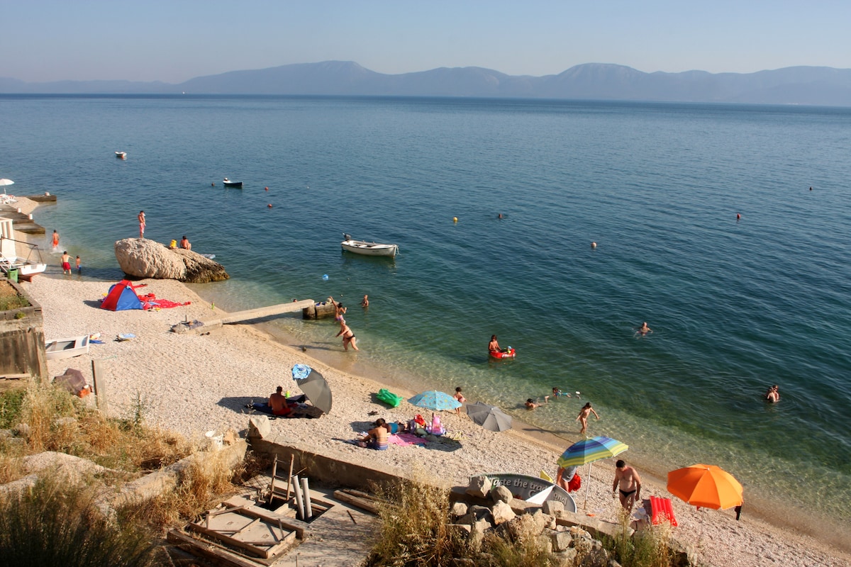 A sandy beach is visible with sunbathers and swimmers enjoying the clear water. Colorful umbrellas provide shade, while small boats can be seen in the distance. The backdrop includes distant mountains under a clear sky, enhancing the serene coastal scene.