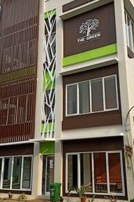 The exterior of a multi-story building is displayed, featuring a blend of wooden panels and green accents. Large windows allow natural light to enter, while a modern logo is prominently placed above the entrance. Surrounding greenery enhances the serene environment.
