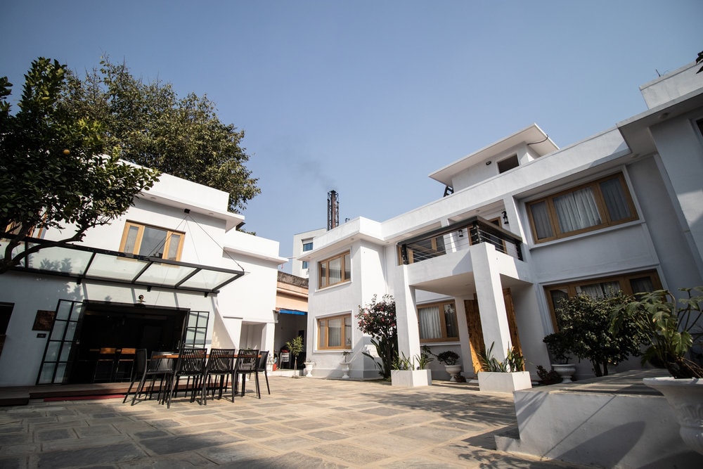 An outdoor view of a white two-story building is captured, surrounded by paved areas and greenery. A separate structure features large glass doors, while a spacious patio hosts a dining table and chairs, inviting gatherings in a bright, open environment under a clear blue sky.