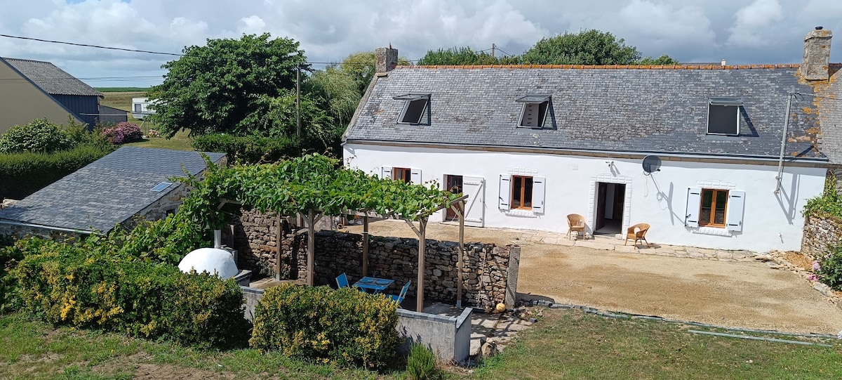 An outdoor view of Ty Gwenn, showcasing the traditional longère structure with a slate roof. A grapevine-covered BBQ terrace is visible, accompanied by outdoor seating. The well-kept garden features colorful greenery, with the spacious farmhouse set against a backdrop of blue sky and scattered clouds.