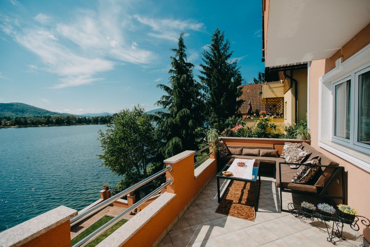 A tranquil balcony overlooks a serene lake, framed by lush trees and distant hills. Outdoor seating is arranged for relaxation, complemented by decorative cushions. A table features refreshments, inviting guests to enjoy the peaceful view in a bright, sunlit atmosphere.