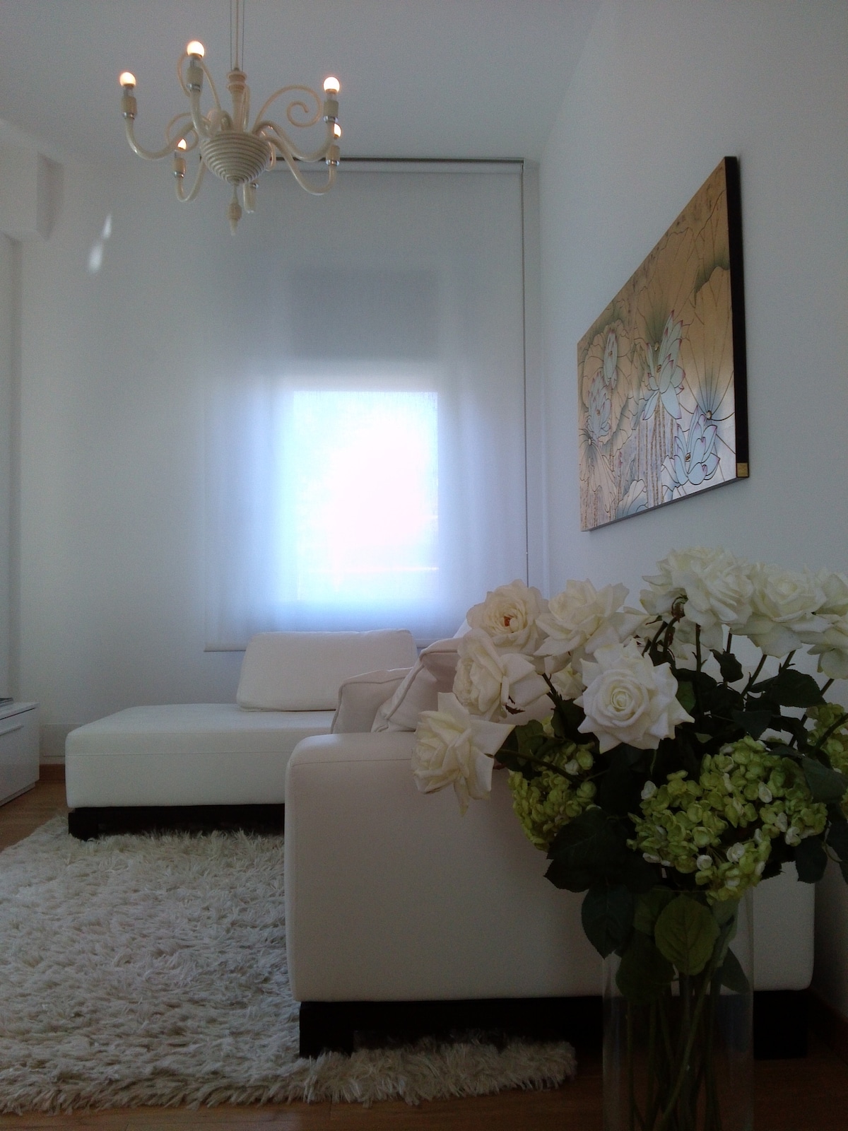 A bright living area features a white sofa set against a soft area rug. A bouquet of white roses and greenery adds a touch of nature. Natural light filters through sheer curtains, illuminating the space. A decorative chandelier hangs above, enhancing the elegant ambiance.