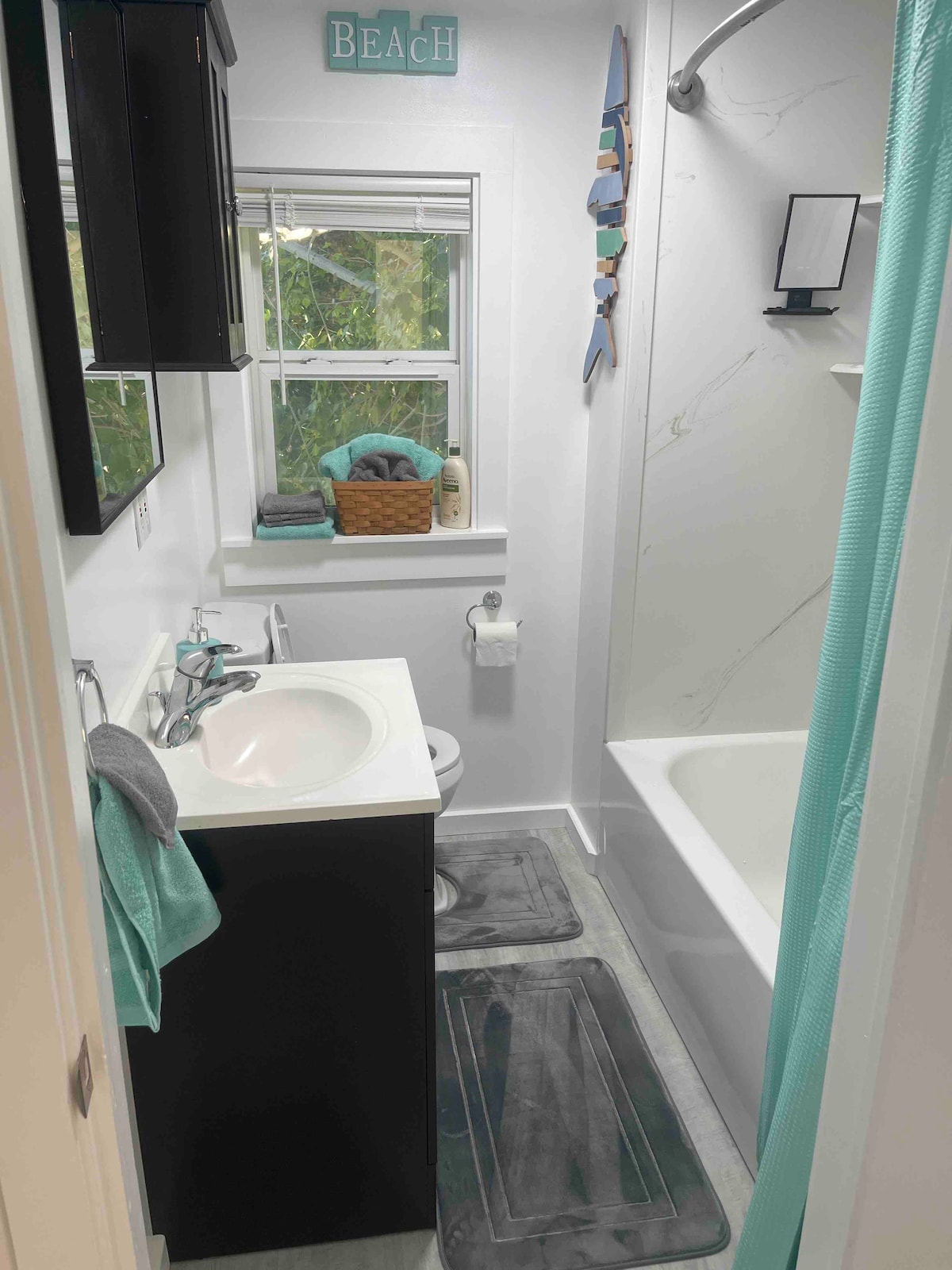 A compact bathroom is shown with a white tub and shower combination. A dark cabinet hosts a sink, while a decorative sign reading 'BEACH' adorns the wall. Soft towels are neatly arranged in a wicker basket near a window, allowing natural light to enter.